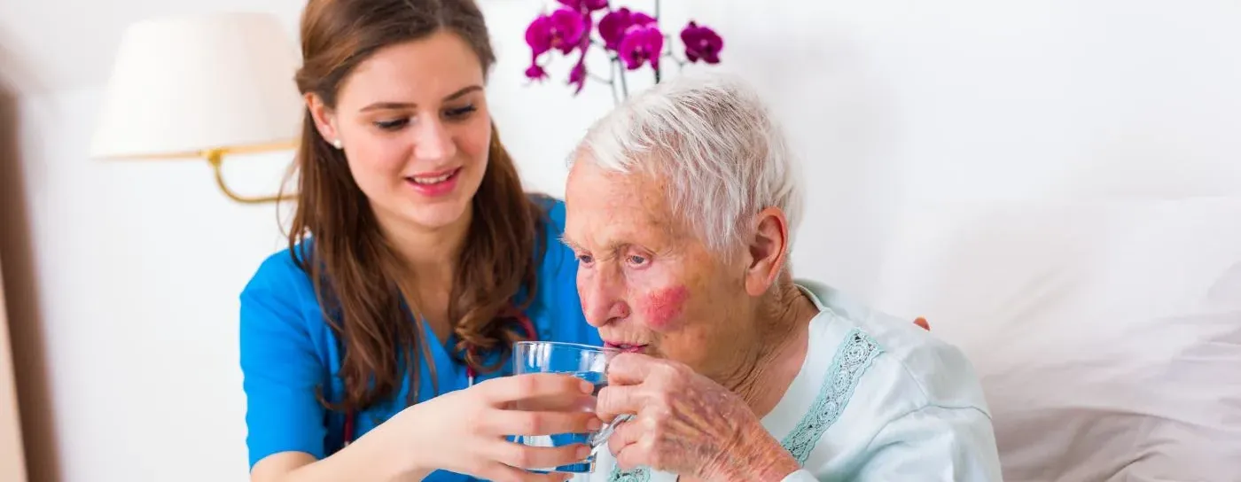 Carer gently helping an older woman in bed to drink as part of end of life care at home in Dartford